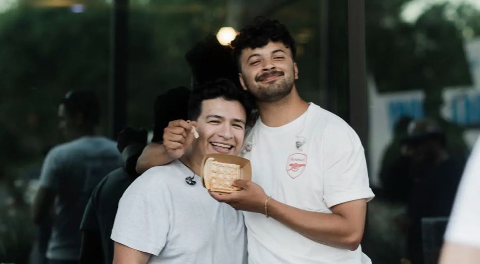 Two young men, one with an arm around the other, grinning at a young adults gathering